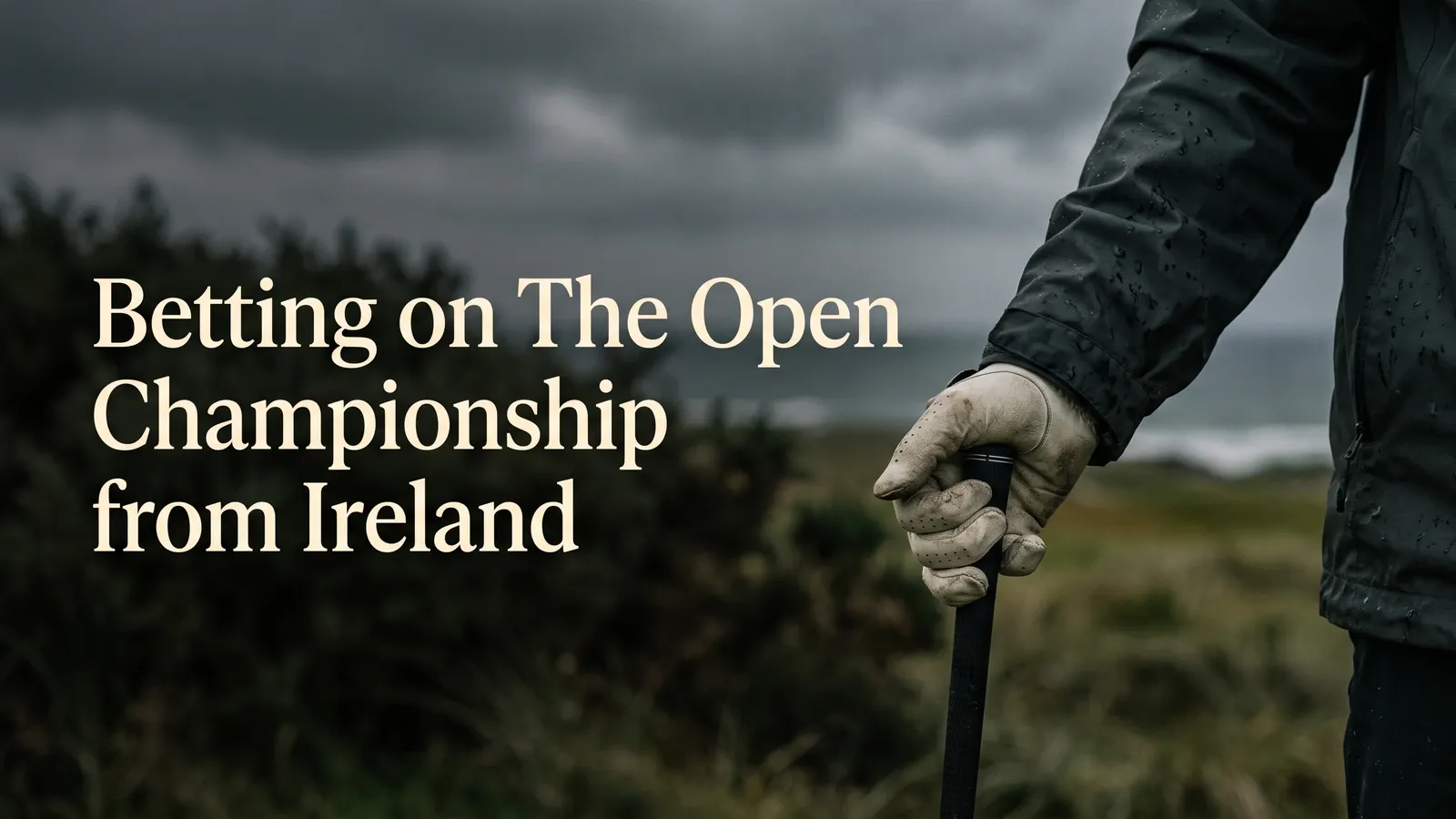 A rain-speckled dark sleeve and worn pale golf glove hold a black club grip against blurred coastal grass and an overcast sky.